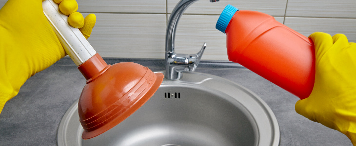 Mãos usando luvas amarelas seguram um desentupidor e um frasco de produto de limpeza sobre uma pia.