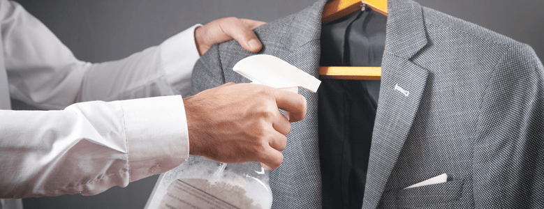 Homem de camisa branca aplicando produto de limpeza com borrifador em um paletó cinza pendurado em um cabide de madeira.